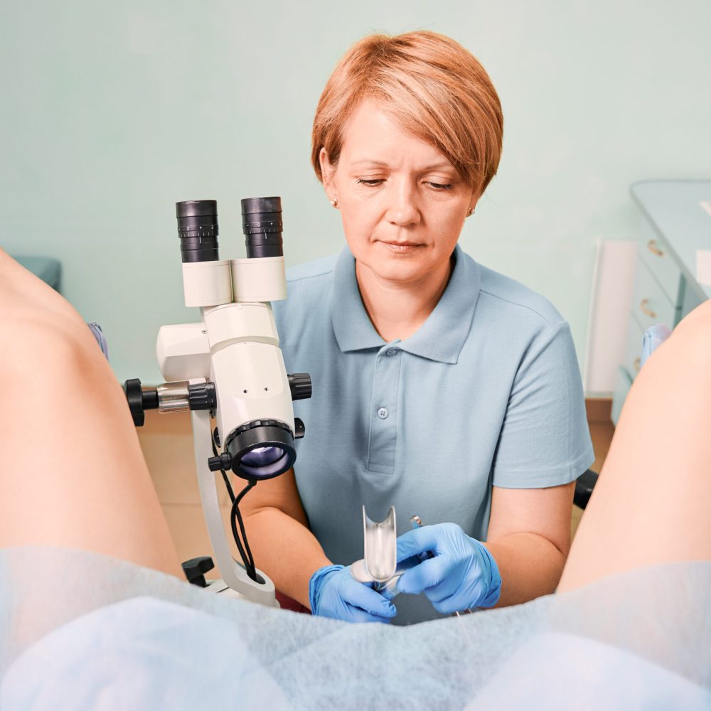 Female gynecologist in sterile gloves using colposcope and gynecological retractors while examining woman in clinic. Patient sitting in gynecological chair. Concept of gynecology and female health.