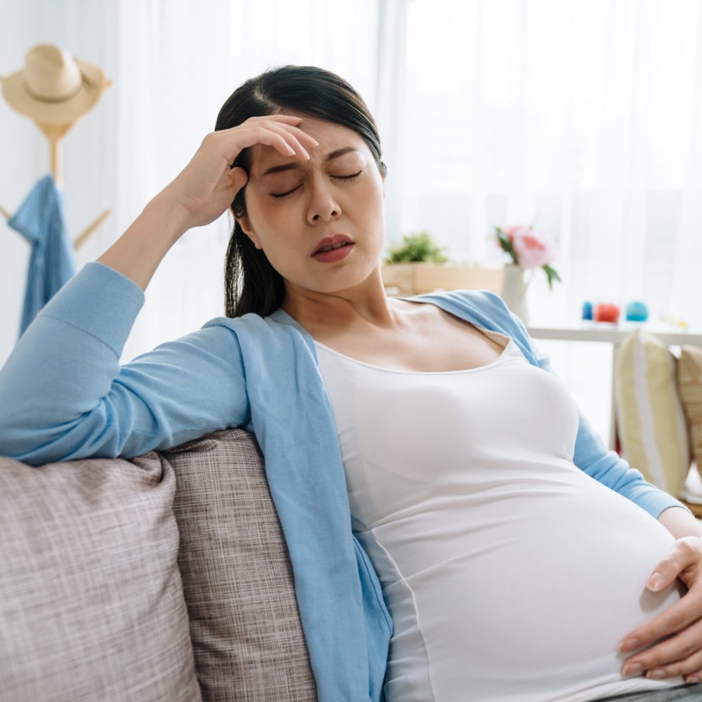 Young asian chinese pregnant woman with headache sitting on sofa couch in living room. Pregnancy symptoms expectation parenthood concept. lady at home holding belly baby feeling hurt at home.